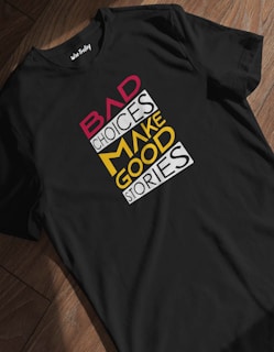 A black t-shirt is laid out on a wooden floor, displaying a bold, colorful quote: 'Bad Choices Make Good Stories.' The words 'Bad' and 'Make' are in red and yellow respectively, while the other words are in white, creating a striking visual contrast.