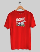 A red T-shirt is hanging on a wooden hanger against a plain background. The front of the shirt features a cartoon panda sleeping on a bamboo branch, accompanied by the words 'BORN TO' with a speech bubble saying 'Zzz...'.