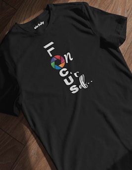 A black T-shirt lies on a wooden floor with the words 'focus on yourself' written vertically. The word 'focus' features a stylized camera shutter icon in multicolor, replacing the first 'o'. The lettering is predominantly white.