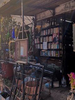 A quaint outdoor shop with a vintage feel, featuring shelves of books and a display of wine bottles on top. There is a mirror hanging on a frame near the entrance, surrounded by plants in clay pots and colorful glass bottles. The setting looks cozy and inviting, with natural and rustic elements.