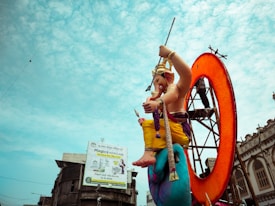 A large, colorful statue of Lord Ganesha seated prominently on a blue pedestal, surrounded by a bright orange circular background. The statue is intricately decorated with garments and jewelry, holding a goad and a loop of rope. The background features a clear blue sky with scattered clouds. A billboard advertising hair care products and an old building with decorative architecture are visible in the mid-ground.
