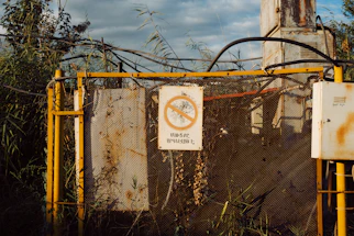 A weathered industrial structure surrounded by overgrown vegetation. The structure is fenced and contains equipment with visible rust and decay. A warning sign with a crossed-out circle and text is prominently displayed.