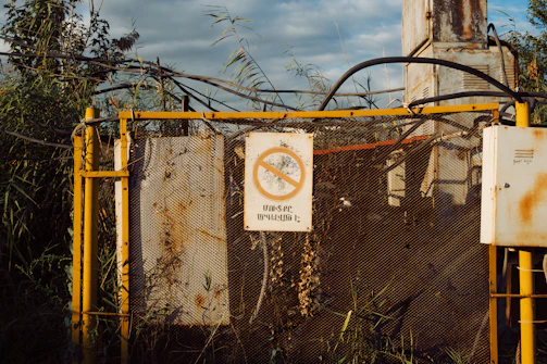 A weathered industrial structure surrounded by overgrown vegetation. The structure is fenced and contains equipment with visible rust and decay. A warning sign with a crossed-out circle and text is prominently displayed.