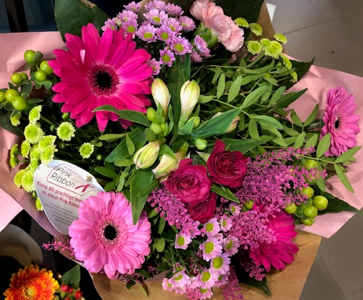 A vibrant bouquet featuring a variety of flowers such as bright pink gerbera daisies, roses, and small chrysanthemums, accented with lush green leaves. A 'Pink Ribbon' label is attached, indicating a support message.