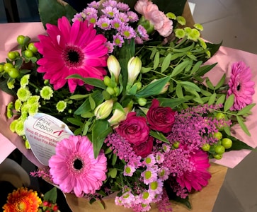 A vibrant bouquet featuring a variety of flowers such as bright pink gerbera daisies, roses, and small chrysanthemums, accented with lush green leaves. A 'Pink Ribbon' label is attached, indicating a support message.