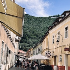 A picturesque street lined with colorful historic buildings and outdoor café seating, bustling with people under a clear blue sky. A green, forested hill rises in the background with a large sign displaying 'BRASOV' atop it.