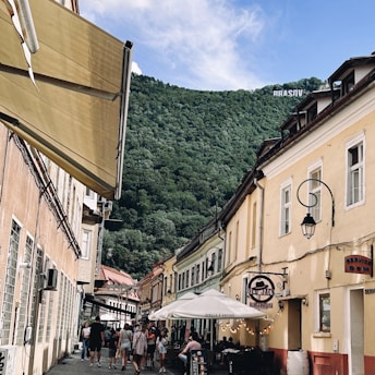 A picturesque street lined with colorful historic buildings and outdoor café seating, bustling with people under a clear blue sky. A green, forested hill rises in the background with a large sign displaying 'BRASOV' atop it.