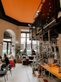 A cozy indoor cafe setting with a high ceiling painted in orange. The cafe has an industrial loft style with metal and wood elements. Several tables are occupied by people socializing and working on laptops. There are plants and homey decorations around, providing a relaxed atmosphere. Large windows allow natural light to flood the space, and some patrons are seated by the windows.