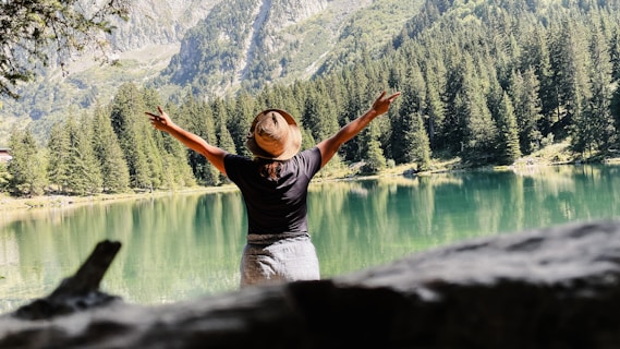 A person wearing a straw hat and dark shirt stands with arms outstretched facing a serene mountain lake surrounded by dense evergreen forest. The background features tall mountains with patches of sunlight creating a peaceful and scenic atmosphere.