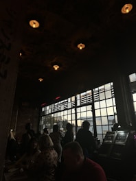 A dimly lit indoor space with a group of people gathered near large industrial-style windows. The ceiling is adorned with exposed bulbs casting a warm glow over the scene. Some people are seated while others stand near the windows, creating silhouettes against the light.