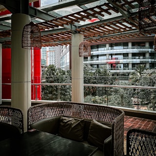 A covered outdoor seating area with wicker-style furniture and cushions is surrounded by urban architecture. The setting includes hanging wicker lamps, tall columns, and a wooden lattice overhead. Glass panels provide a view of nearby buildings and lush green trees, creating a blend of natural and urban elements.