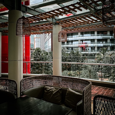A covered outdoor seating area with wicker-style furniture and cushions is surrounded by urban architecture. The setting includes hanging wicker lamps, tall columns, and a wooden lattice overhead. Glass panels provide a view of nearby buildings and lush green trees, creating a blend of natural and urban elements.