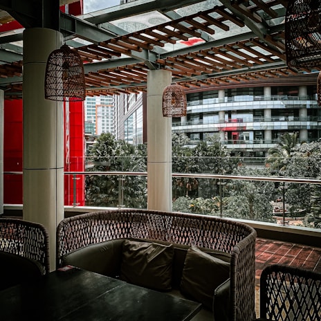 A covered outdoor seating area with wicker-style furniture and cushions is surrounded by urban architecture. The setting includes hanging wicker lamps, tall columns, and a wooden lattice overhead. Glass panels provide a view of nearby buildings and lush green trees, creating a blend of natural and urban elements.