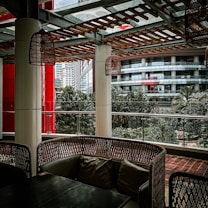 A covered outdoor seating area with wicker-style furniture and cushions is surrounded by urban architecture. The setting includes hanging wicker lamps, tall columns, and a wooden lattice overhead. Glass panels provide a view of nearby buildings and lush green trees, creating a blend of natural and urban elements.