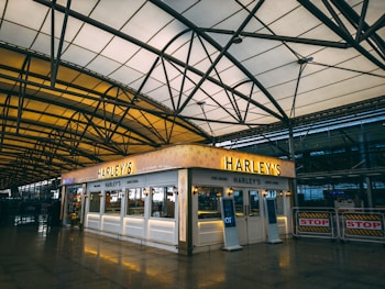 A caf&eacute; named Harley's positioned in what appears to be an airport terminal. The caf&eacute; features a bright sign and a well-lit display with pastries and beverages visible. The architecture showcases a spacious, well-designed arched ceiling with a modern aesthetic.