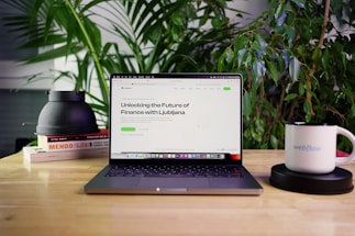 A laptop displaying a webpage titled 'Unlocking the Future of Finance with Ljubljana' is positioned on a wooden table. Surrounding the laptop are green plants, a stack of books, a black object, and a white mug with the word 'webflow' printed on it. The setting appears to be a comfortable workspace or home office.