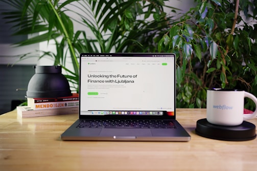 A laptop displaying a webpage titled 'Unlocking the Future of Finance with Ljubljana' is positioned on a wooden table. Surrounding the laptop are green plants, a stack of books, a black object, and a white mug with the word 'webflow' printed on it. The setting appears to be a comfortable workspace or home office.