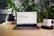 A laptop displaying a webpage titled 'Unlocking the Future of Finance with Ljubljana' is positioned on a wooden table. Surrounding the laptop are green plants, a stack of books, a black object, and a white mug with the word 'webflow' printed on it. The setting appears to be a comfortable workspace or home office.