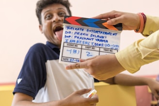 A person is playfully sticking out their tongue while looking at the camera. In front of them, someone is holding a movie clapperboard with colorful stripes and text indicating it's for a film scene. The clapperboard displays information such as director and camera details. The setting appears casual.