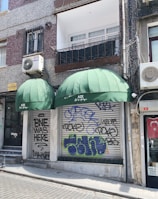 A storefront with two large green awnings, one of which displays the name 'AZE Art & Antique.' Graffiti covers the metal shutters below the awnings, with various tags and phrases. The building features a mix of texture with speckled and smooth surfaces. An air conditioning unit is mounted on the wall above, and a Turkish flag is displayed to the side.