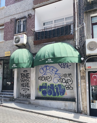 A storefront with two large green awnings, one of which displays the name 'AZE Art & Antique.' Graffiti covers the metal shutters below the awnings, with various tags and phrases. The building features a mix of texture with speckled and smooth surfaces. An air conditioning unit is mounted on the wall above, and a Turkish flag is displayed to the side.