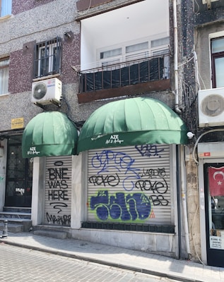 A storefront with two large green awnings, one of which displays the name 'AZE Art & Antique.' Graffiti covers the metal shutters below the awnings, with various tags and phrases. The building features a mix of texture with speckled and smooth surfaces. An air conditioning unit is mounted on the wall above, and a Turkish flag is displayed to the side.