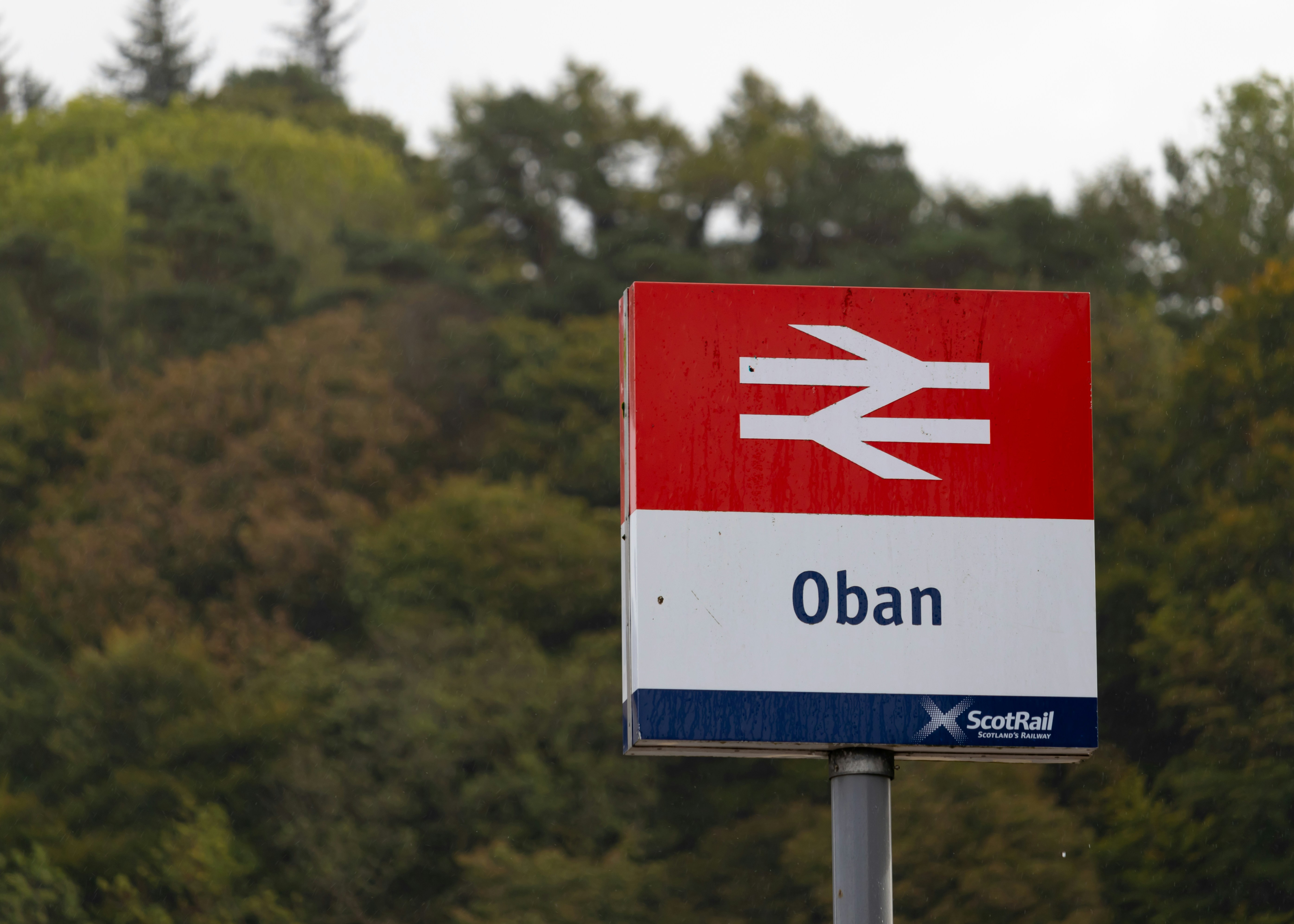 a red white and blue sign and some trees, Oban Train Station Sign