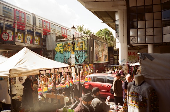 A bustling outdoor market scene with vendors selling colorful merchandise under tents. A red train passes on an elevated track above, and vibrant street art decorates the surrounding walls. Several people are browsing stalls, some with clothing and crafts displayed.