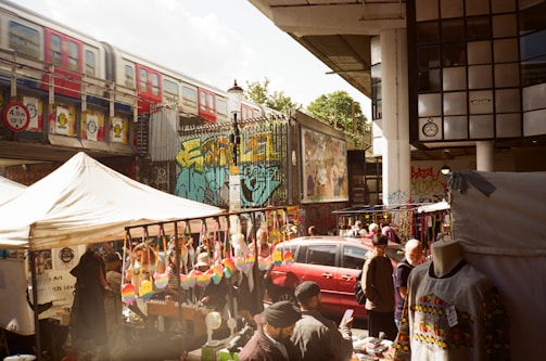 A bustling outdoor market scene with vendors selling colorful merchandise under tents. A red train passes on an elevated track above, and vibrant street art decorates the surrounding walls. Several people are browsing stalls, some with clothing and crafts displayed.