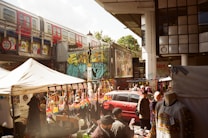 A bustling outdoor market scene with vendors selling colorful merchandise under tents. A red train passes on an elevated track above, and vibrant street art decorates the surrounding walls. Several people are browsing stalls, some with clothing and crafts displayed.
