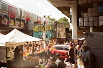 A bustling outdoor market scene with vendors selling colorful merchandise under tents. A red train passes on an elevated track above, and vibrant street art decorates the surrounding walls. Several people are browsing stalls, some with clothing and crafts displayed.