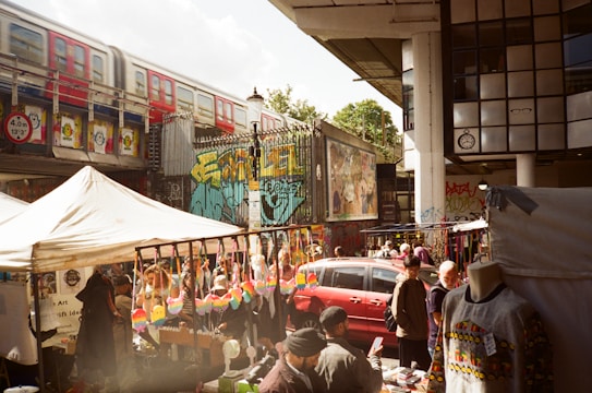 A bustling outdoor market scene with vendors selling colorful merchandise under tents. A red train passes on an elevated track above, and vibrant street art decorates the surrounding walls. Several people are browsing stalls, some with clothing and crafts displayed.