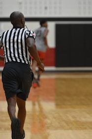 A basketball referee in motion on a court, with black shorts and a black and white striped shirt. The background features another person in athletic wear, blurred, along with a gymnasium setting.