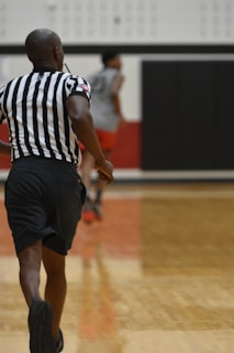 A basketball referee in motion on a court, with black shorts and a black and white striped shirt. The background features another person in athletic wear, blurred, along with a gymnasium setting.