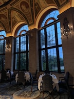 An elegant interior space featuring large arched windows and high ceilings adorned with classical paintings. The dining area includes tables with white tablecloths surrounded by ornate chairs, while elaborate chandeliers add a touch of sophistication. The view outside shows twilight skies and silhouettes of palm trees.
