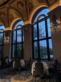 An elegant interior space featuring large arched windows and high ceilings adorned with classical paintings. The dining area includes tables with white tablecloths surrounded by ornate chairs, while elaborate chandeliers add a touch of sophistication. The view outside shows twilight skies and silhouettes of palm trees.