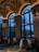 An elegant interior space featuring large arched windows and high ceilings adorned with classical paintings. The dining area includes tables with white tablecloths surrounded by ornate chairs, while elaborate chandeliers add a touch of sophistication. The view outside shows twilight skies and silhouettes of palm trees.