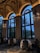 An elegant interior space featuring large arched windows and high ceilings adorned with classical paintings. The dining area includes tables with white tablecloths surrounded by ornate chairs, while elaborate chandeliers add a touch of sophistication. The view outside shows twilight skies and silhouettes of palm trees.
