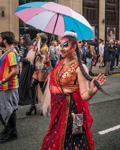 A vibrant and colorful street event with diverse participants. A person dressed in ornate, bright clothing holds a multicolored umbrella, smiling and interacting with the crowd. The setting appears urban, with people wearing a variety of creative costumes walking along a street, and many onlookers in the background.