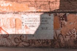 A worn brick wall with a large poster displaying Spanish text. The wall is weathered with various shades of orange and brown, and there is graffiti surrounding the poster. Shadows fall across the wall, indicating sunlight from above.