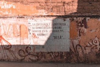 A worn brick wall with a large poster displaying Spanish text. The wall is weathered with various shades of orange and brown, and there is graffiti surrounding the poster. Shadows fall across the wall, indicating sunlight from above.