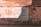 A worn brick wall with a large poster displaying Spanish text. The wall is weathered with various shades of orange and brown, and there is graffiti surrounding the poster. Shadows fall across the wall, indicating sunlight from above.