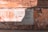 A worn brick wall with a large poster displaying Spanish text. The wall is weathered with various shades of orange and brown, and there is graffiti surrounding the poster. Shadows fall across the wall, indicating sunlight from above.