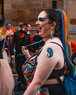 A person with a colorful rainbow ponytail and bold makeup is laughing joyfully. They are wearing a sequined outfit with a yin-yang tattoo visible on their arm. The background features other colorfully dressed individuals, suggesting a festive outdoor event.