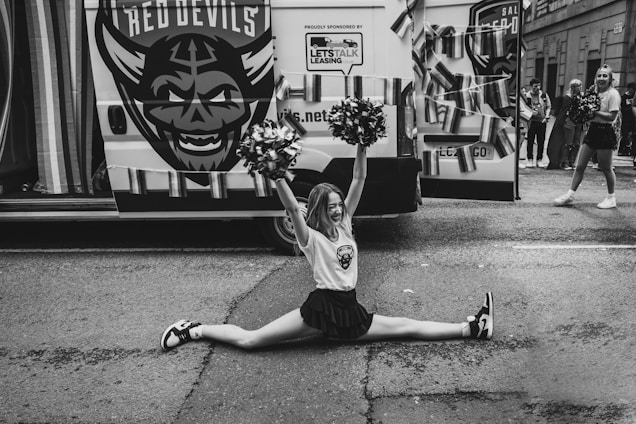 A cheerleader performs a split in front of a van decorated with a large logo and streamers. She holds pom-poms and smiles while other cheerleaders and people in the background can be seen. The setting appears to be an outdoor event or parade.