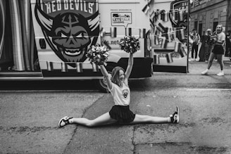A cheerleader performs a split in front of a van decorated with a large logo and streamers. She holds pom-poms and smiles while other cheerleaders and people in the background can be seen. The setting appears to be an outdoor event or parade.