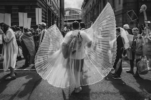 A group of people gathering on a city street, participating in an event or parade. The focal point is an individual wearing large, translucent wing-like fabric creating a visual centerpiece. Other participants interact, holding signs and carrying various items while the background showcases urban architecture.