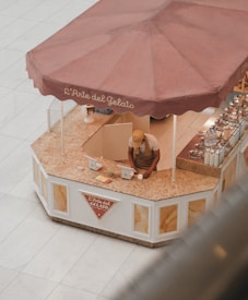 A person wearing a cap and apron is working at a gelato kiosk with a large maroon umbrella and the text 'L'Arte del Gelato' on it. The kiosk counter appears to be made of marble, and there are two point-of-sale devices on the counter. The kiosk is situated in an open space with a tiled floor.