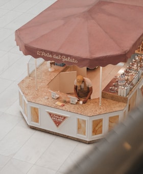 A person wearing a cap and apron is working at a gelato kiosk with a large maroon umbrella and the text 'L'Arte del Gelato' on it. The kiosk counter appears to be made of marble, and there are two point-of-sale devices on the counter. The kiosk is situated in an open space with a tiled floor.