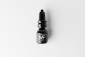 A black nasal spray bottle with a nozzle lid is placed against a light background. The label on the bottle features large, bold letters 'MF'. The design is sleek and minimalist, conveying a modern aesthetic.